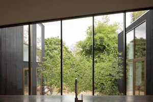Modern kitchen interior featuring a dark countertop, matte black faucet, and large panoramic windows overlooking a dense green forest with dark vertical wood cladding on the exterior walls.