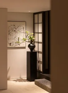 Modern minimalist entryway featuring a dark wood pedestal with an ikebana floral arrangement, abstract ink painting on the wall, and a black shoji screen under warm lighting.