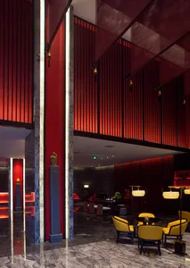 Luxurious hotel lobby interior with red marble columns, dramatic red slatted walls, yellow upholstered armchairs, and warm pendant lighting in a double-height space.