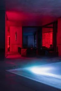 Modern indoor swimming pool surrounded by concrete architecture, featuring modular dark lounge seating, dramatic red uplighting on the ceiling and walls, and bright blue underwater lighting in the pool.