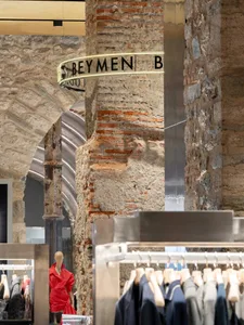 Interior view of a fashion boutique featuring rough exposed brick walls, stone arches, modern stainless steel clothing racks, and a mannequin wearing a bright red dress.