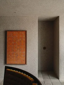 Modern entryway featuring grey terrazzo walls and floor, a large framed orange geometric tile artwork, and the back of a curved mustard-velvet chair in the foreground.