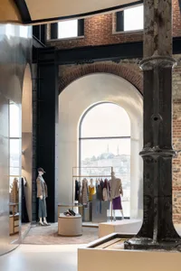 Interior view of a fashion boutique featuring large arched windows, exposed brick walls, heavy cast-iron columns, minimalist clothing displays, and a panoramic city view of Istanbul with a mosque dome in the distance.