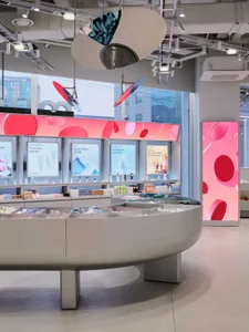 Modern beauty store interior with a large curved grey counter, colorful product displays, digital pink LED wall graphics, and hanging sculptural light fixtures.