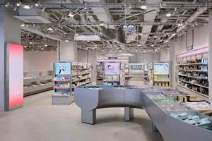 Interior view of a modern cosmetics and skincare store featuring a large curved grey display island, product shelving, digital advertisements, and exposed ceiling infrastructure.