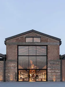 Front exterior view of a luxury retail store with a large black-framed glass facade set within a restored red brick and stone industrial building at dusk.