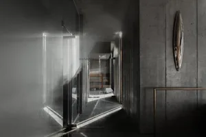 Narrow brutalist concrete hallway featuring a large translucent glass partition wall, dramatic diagonal light beams, and an asymmetrical metallic wall sculpture.