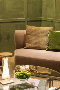 Close-up of a modern living room corner with a taupe velvet sofa, mustard patterned pillow, green velvet pillow, and an olive green textured wall panel background. A glass coffee table in the foreground holds a bowl of green grapes and a marble lamp.