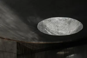 View looking up at a large circular concrete oculus skylight set into a dark raw concrete ceiling with hanging chainmail curtain below.