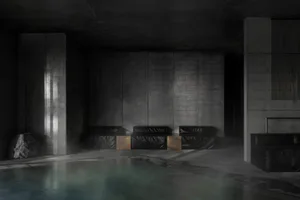 A dimly lit brutalist indoor swimming pool surrounded by massive concrete walls, featuring black leather modular benches with bronze accents and a large natural boulder on the left.