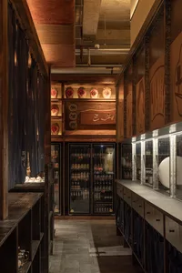 Narrow interior corridor of a Japanese izakaya featuring reclaimed wood shelves, hanging indigo noren curtains, illuminated ceramic sake jars, and glass-door refrigerators stocked with beverages.