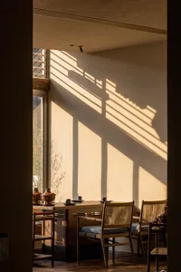 A tranquil wabi-sabi style dining room featuring wooden table and chairs with woven rattan backs, illuminated by warm sunlight that creates strong linear shadows on a textured plaster wall.