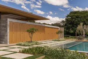 Modern outdoor pool area with a long light-wood perforated screen wall, concrete structure, stone pillars, stepping stone pathway, and lush tropical landscaping under a bright blue sky.
