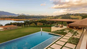 Modern hillside villa with a long rectangular infinity pool overlooking a scenic mountain lake and forested hills at sunset, with a woman standing at the pool edge.