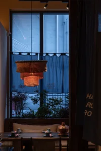 Intimate restaurant dining area featuring a large three-tiered handmade linen pendant lamp glowing warmly in front of a tall window with blue noren curtain at dusk.