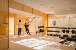 Modern home gym interior with bright orange perforated metal room dividers, two people training with suspension straps, dumbbell racks, and strong natural sunlight casting long shadows on the floor.
