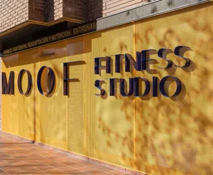 Exterior view of MOOF Fitness Studio featuring large 3D blue lettering mounted on a bright yellow perforated metal wall with terracotta tile flooring.