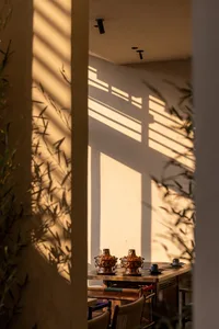 A wooden dining table set with two copper hot pots, viewed through a doorway with dramatic bamboo leaf shadows cast by warm sunlight on the wall.
