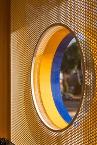 Close-up of a circular opening with yellow and blue painted rings set within a golden perforated metal screen, with trees visible in the background.