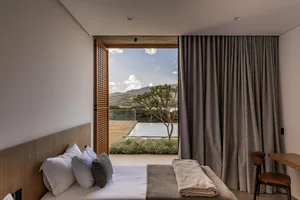 Modern minimalist hotel bedroom with light wood headboard, white and grey bedding, and large open doors leading to a terrace with mountain and pool view.