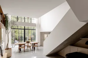 Modern double-height dining room featuring a large wooden table, woven dining chairs, concrete staircase, and expansive black-framed windows overlooking a garden.