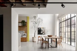 Modern industrial loft dining room featuring a long oak dining table with mixed wooden chairs, a large potted tree, white brick walls, and large black-framed windows allowing abundant natural light.
