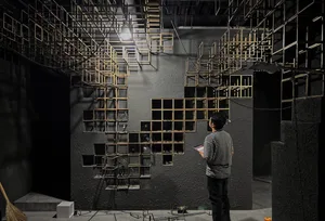 Man in striped shirt observing a large-scale wooden cube grid installation mounted on a dark concrete wall in an industrial exhibition space with dramatic overhead lighting.