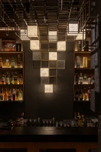Luxurious bar interior featuring a dramatic textured dark stone wall with an illuminated onyx and brass grid installation, flanked by backlit liquor shelves filled with premium bottles and glassware.