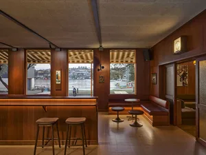Mid-century modern cafe interior featuring a long teak wood bar, leather banquette seating, round pedestal tables, and large windows overlooking a harbor with boats and distant hills.