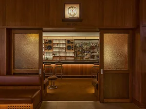 View through textured glass doors into a mid-century modern home bar featuring walnut wood paneling, leather seating, bar stools, and an extensive vinyl record collection.
