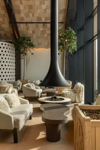 Modern lounge interior featuring a tall sculptural black metal fireplace, curved wooden coffee tables, cream sofas with fluffy sheepskin throws, potted trees, and light wood block walls under a high ceiling.