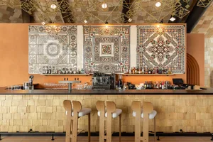 Modern bar counter with warm wood block paneling, four wooden bar stools, colorful patterned terracotta tiles on the back wall, and geometric brass pendant lights hanging from the ceiling.