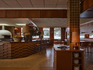 Interior view of a retro-style restaurant with a curved terracotta brick bar, wood-fired pizza oven, warm wood paneling, and leather bar stools under soft lighting.