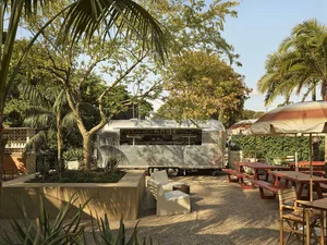 Outdoor tropical garden lounge with a vintage silver Airstream trailer converted into a bar, surrounded by palm trees, concrete planters with aloe and succulents, wooden picnic tables, and modern outdoor seating under a large striped umbrella.