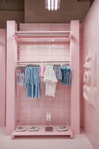 Pink tiled retail display alcove with clothing racks holding pastel and striped garments, white ballet flats on a pink platform, and a large sculptural pink face on the right wall.