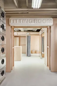 View into a modern minimalist fitting room area with light wood frames, numbered booths 01, 02, 03, grey curtains, and a white sign reading FITTING ROOM.
