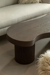 Close-up view of a dark walnut coffee table with an organic, kidney-bean shape and cylindrical pedestal base, placed on a light tile floor in front of a cream-colored sofa with a fluffy throw pillow.