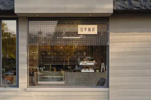 Street view of a modern minimalist coffee shop with textured concrete walls, a large glass window revealing the warm-lit interior, wooden signage reading 四季咖啡, and a metal mesh screen.