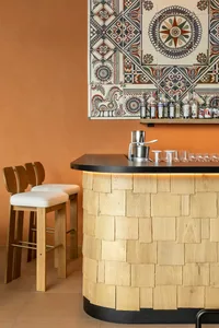 Curved home bar with light wood shingle cladding, black countertop, under-counter lighting, three wooden bar stools with white cushions, and a vibrant patterned tile wall featuring geometric and folk motifs against a warm terracotta background.