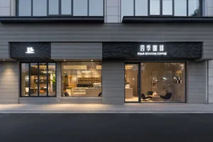 Street view of Four Seasons Coffee shop featuring a modern grey textured concrete facade, large glass windows revealing the interior, and illuminated black stone signage at dusk.