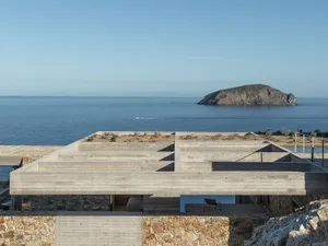 Modern exposed concrete rooftop terrace overlooking the deep blue Aegean Sea with a small boat and rocky island in the distance.