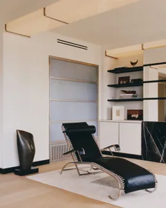 Modern minimalist living room featuring a black leather and chrome chaise lounge on a white rug, with built-in shelving, marble accent wall, and abstract bronze sculpture.
