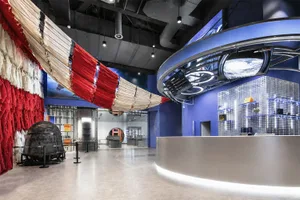 Interior view of a modern science museum exhibition hall featuring a large red and white parachute canopy installation hanging from the ceiling, a curved metallic reception desk with LED lighting, blue walls with digital space displays, and a vintage space capsule on the left.
