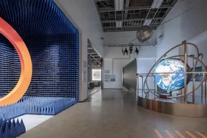 Interior view of a science museum exhibition space featuring a large interactive globe on a metallic armature, a massive blue acoustic foam wall with an orange circular accent, and a hanging satellite model under industrial ceiling infrastructure.