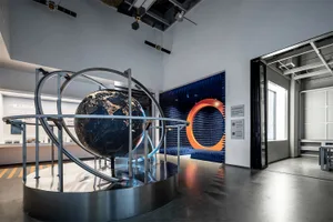 Large metallic globe exhibit with orbital rings displayed on a circular platform inside a contemporary science museum with blue acoustic panels and industrial ceiling.