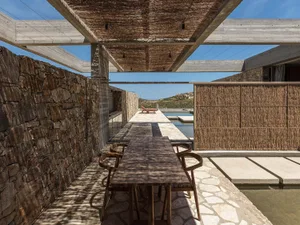 Outdoor dining area in a modern Mediterranean courtyard featuring rough stone walls, a reed pergola, wooden dining table and chairs, and a narrow reflecting pool under a clear blue sky.