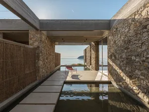 Modern villa courtyard with stone walls, concrete ceiling, water channel, red chair, and sea view beyond
