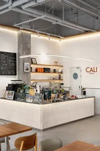 Modern minimalist cafe interior featuring a light wood and concrete service counter, white tiled walls, exposed ceiling pipes, and warm pendant lighting.
