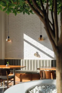 Modern cafe interior featuring white painted brick wall, built-in upholstered booth, wooden tables and chairs, and pendant lights with strong natural sunlight creating sharp shadows.