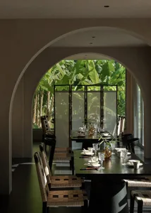 Modern dining room viewed through a large arched opening, featuring a long dark wood table set with white ceramics and crystal glasses, surrounded by black-framed chairs with woven rattan seats, with lush green banana plants visible in the garden beyond.